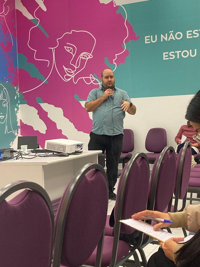 Alcyr Barbin Neto dando palestra com microfone na mão e painel lilás com desenho de mulher ao fundo.